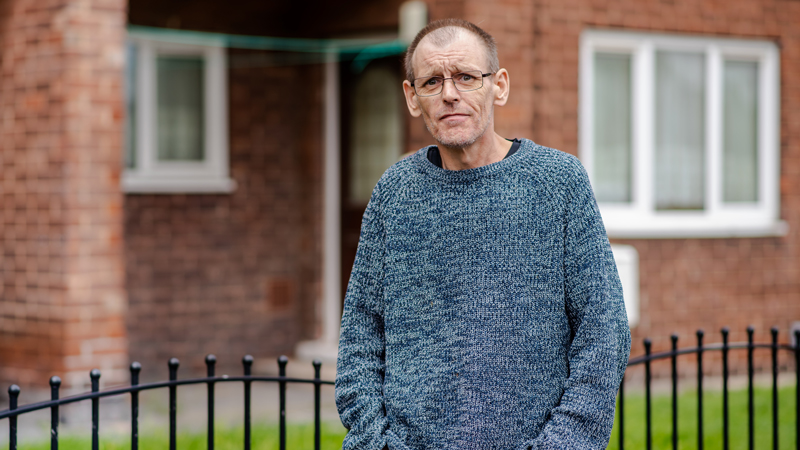 Lee standing in front of a house. He has his hands in his pockets,