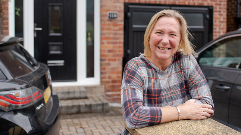 Saragh standing outside a house. She is leaning against a wall. There are two cars at the edge of the image.